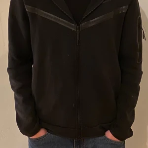 Nike tech fleece - Svart Nike tech fleece som är ungefär 1 år gammal men har mycket kvar att ge. Jag köpte den på JD hemsida för 1300kr och säljer den nu för bara 599kr eftersom att den är andvänd. Den är i toppskick och har inga skavanker.