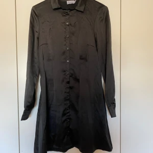 Svart klänning - Svart klänning med knappar framtill i glansigt material. Långa ärmar och krage. Storlek 36 från NAKD