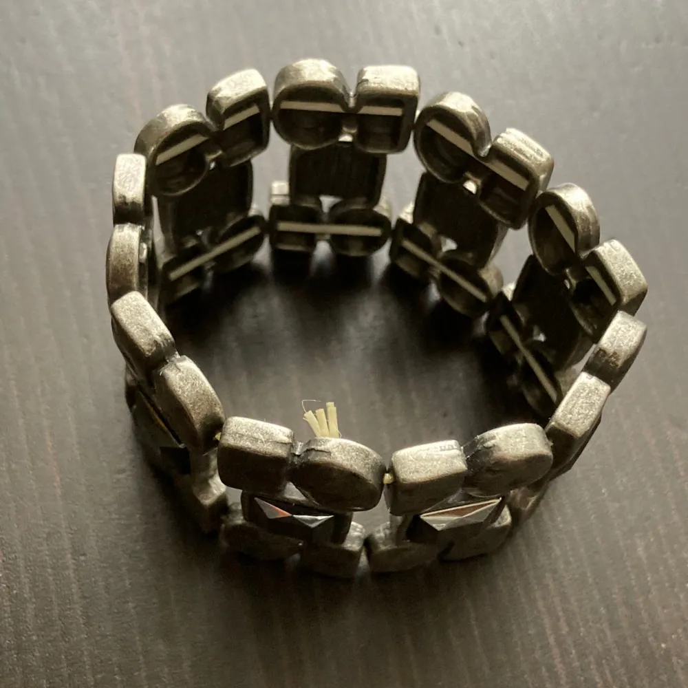 Elastiskt armband i silverfärgad metall med glansiga stenar. Har kortats av. Bredd: 3,9 cm, innerdiameter: 4,6 cm.. Asusteet.