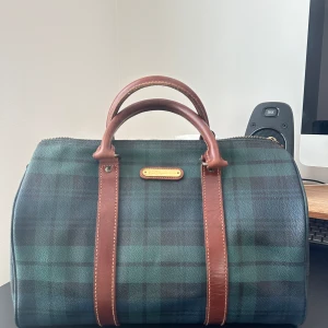Polo Ralph Lauren Boston - Polo Ralph Laurens klassiska Boston väskan som kombinerar elegans och funktionalitet! Den mörkgröna canvasen med bruna läderdetaljer är verkligen ett tidlöst signum för varumärket. Denna typ av design är inte bara praktisk utan också en perfekt representation av Polo Ralph Laurens ikoniska preppy-stil och förkärlek för traditionellt amerikanskt mode.  Väskan är väl omhändertagen och har fortfarande många års användning kvar. Perfekt för någon som söker en stilren och praktisk accessoar.
