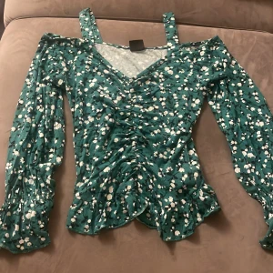 somrig grön topp - en grön topp med vita blommor som passar perfekt inför sommaren. Armarna och slutet på toppen är flared/ruffled. Tröjan är i bra skick men använd x antal gånger (pris kan diskuteras vid snabba köp!!)
