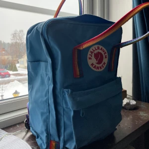 Fjällräven kånken ljusblå rainbow - Knappt använd fjällräven ryggsäck. Har stått orörd många år. Väskan har fack på sidorna för vattenflaska, även ett fack inuti där ett medhavt sittunderlag finns. Axelband går att justera