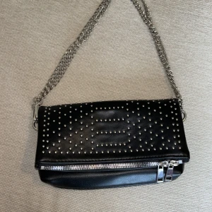 Svart axelremsväska nitar  - Liten väska med nit-detaljer. Axelbandet är 110 cm lång men kan även träs dubbelt i ”öglorna” så som på bilden. H: 13 cm. L: 23cm.