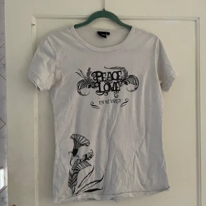 Vit t-shirt med tryck från Carlings Merch - Säljer en vit t-shirt från Carlings Merch i storlek L. T-shirten har ett coolt tryck på framsidan med texten 'Peace & Love En Ny Värld' och ett blommönster. På baksidan finns en lista med artistnamn som Rihanna, Regina Spektor, Kent och många fler. Perfekt för festivaler eller vardagsbruk. T-shirten är i bra skick och gjord av bomull. Passa på att köpa denna unika t-shirt!