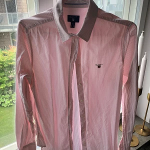 Rosa randig skjorta från GANT - En stilren rosa randig skjorta från GANT med en klassisk krage och långa ärmar. Skjortan har knappar framtill och en liten broderad logotyp på bröstet.