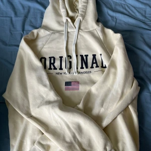 Beige hoodie med texten 'ORIGINAL' - En beige hoodie från ginateicot med texten 'ORIGINAL' i mörkblått och en liten amerikansk flagga under. Hoodien har en justerbar huva med snören och långa ärmar.
