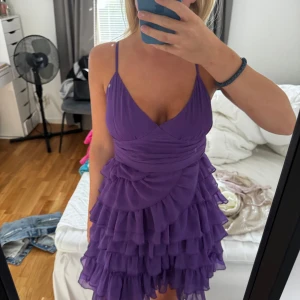 Lila klänning med volanger - En lila klänning med volanger och smala axelband. Klänningen har en omlott-design framtill och en öppen rygg. Perfekt för festliga tillfällen.