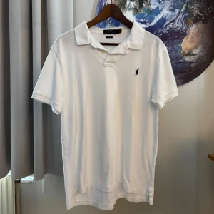 Vit kortärmad t-shirt från Polo Ralph Lauren - Säljer en snygg vit kortärmad t-shirt från Polo Ralph Lauren. Den är i slim fit och har en klassisk krage med två knappar. Materialet är bomull och den har en liten broderad logga på bröstet. Perfekt för både vardag och fest!