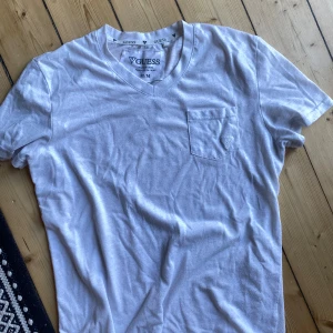 Vit t-shirt från Guess - Säljer en snygg vit t-shirt från Guess i storlek M. T-shirten har en v-ringning och en liten bröstficka med Guess-loggan broderad på. Perfekt för en avslappnad och stilren look. Passar bra till både vardag och fest!