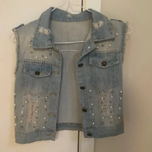 Ljusblå jeansväst med pärlor och slitningar - Säljer en snygg ljusblå jeansväst i storlek M. Västen har coola slitningar och är dekorerad med pärlor på framsidan. Den har två bröstfickor med knappar och en klassisk krage. Perfekt för en avslappnad och trendig look! st beroende på hur man vill den ska sitta