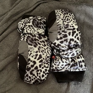 Svartvita leopardmönstrade tumvantar - Ett par svartvita leopardmönstrade tumvantar med förstärkta svarta partier på handflatan och tummen. Vantarna har elastiska muddar och justerbara remmar för bättre passform. med Flis material och fingrarna har som ett eget rum typ. använd 1 skidsemester 