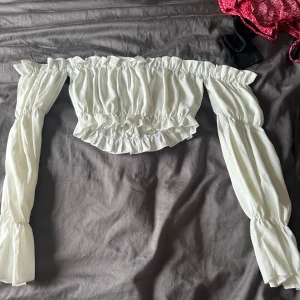 Vit off-shoulder topp - En vit off-shoulder topp med långa ärmar och elastiska detaljer vid axlar, ärmslut och midja. Toppen har en rynkad design och är croppad.