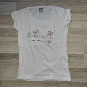 Vit t-shirt med fjärilar och text - En vit t-shirt med korta ärmar och ett dekorativt mönster av fjärilar och text på framsidan. T-shirten är gjord av bomull och har en normal passform. stl 146/152