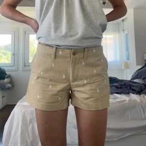 Beige shorts från Ralph Lauren Sport - Beige shorts från Ralph Lauren Sport med broderade vita hästar. Shortsen har fickor fram och bak samt en knapp och dragkedja i midjan.