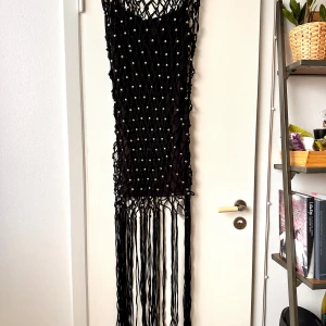 Svart klänning med pärlor och fransar - En svart klänning dekorerad med pärlor och långa fransar. Klänningen har ett nätliknande mönster och är ärmlös. Aldrig använd! 