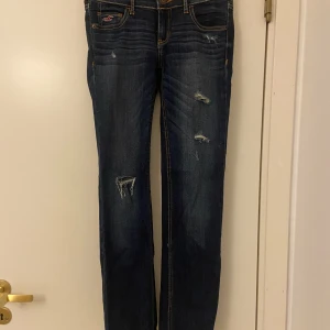Hollister lågmidjade jeans, stl 24/31 - Mörkblå lågmidjade jeans med hål över knät och fickorna i fint skick.  Frakt tillkommer, men kläder som köps kan samfraktas!