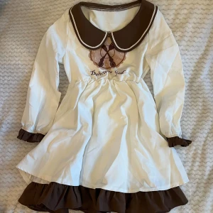 Vit och brun klänning med Peter Pan-krage - En söt vit och brun klänning med långärmad design och en Peter Pan-krage. Klänningen har en volangdetalj vid ärmslut och nederkant samt ett tryck på framsidan. Köpt på Kawaii Shop!