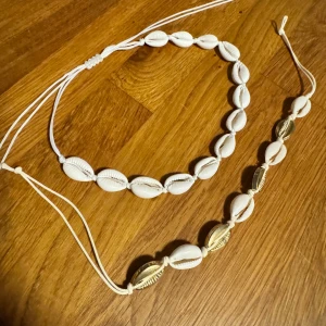 Halsband och armband med snäckor - Ett set bestående av ett halsband och ett armband, båda dekorerade med vita snäckor. Halsbandet har en justerbar rem och är helt gjort av vita snäckor. Armbandet har en kombination av vita och guldfärgade snäckor, även det med en justerbar rem. Halsbandet är använt en gång. Armbandet aldrig använt. 