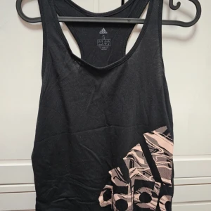 Svart linne från Adidas - Ett svart linne från Adidas med ett stort rosa mönster på framsidan. Linnet har en rund halsringning och är ärmlöst. Tillverkat i Climalite-material för att hålla dig torr och bekväm.