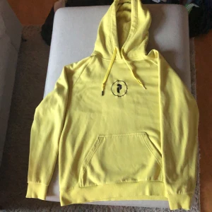 Gul hoodie från Peak Performance - En gul hoodie från Peak Performance med svart logotyp på bröstet och ryggen. Hoodien har en känguruficka framtill och justerbar huva med dragsko. Ribbstickade muddar vid ärmslut och nederkant.