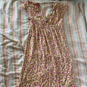 Blommig klänning från Monki - En somrig klänning från Monki med ett blommigt mönster i rosa och grönt på en gul bakgrund. Klänningen har en v-ringad hals och volangdetaljer. Den är ärmlös och har en elastisk midja för en bekväm passform.