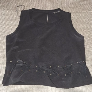 Svart ärmlös topp med snörning - En svart ärmlös topp med snörningsdetaljer vid midjan. Toppen har en enkel design med rund halsringning och är tillverkad i ett mjukt material. använt den bara 1 fång på min födelsedag och sen det var inte längre på min storlek 🥲🥲🥲