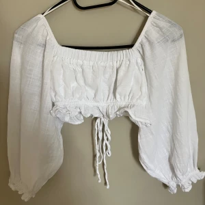 Vit crop-top med långa puffärmar  - Mått: XS. Skick: Nyskick. Fluffig och supersjuk topp med djup, fyrkantig halsringning. Ryggen är öppen med justerbara snören i samma tyg som toppen. 