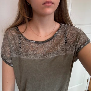 Detaljerad T-shirt  - Detaljerad T-shirt i khaki färg. Spets upptill och pärlor/stenar runt ärmarna och halsringningen. Storlek M
