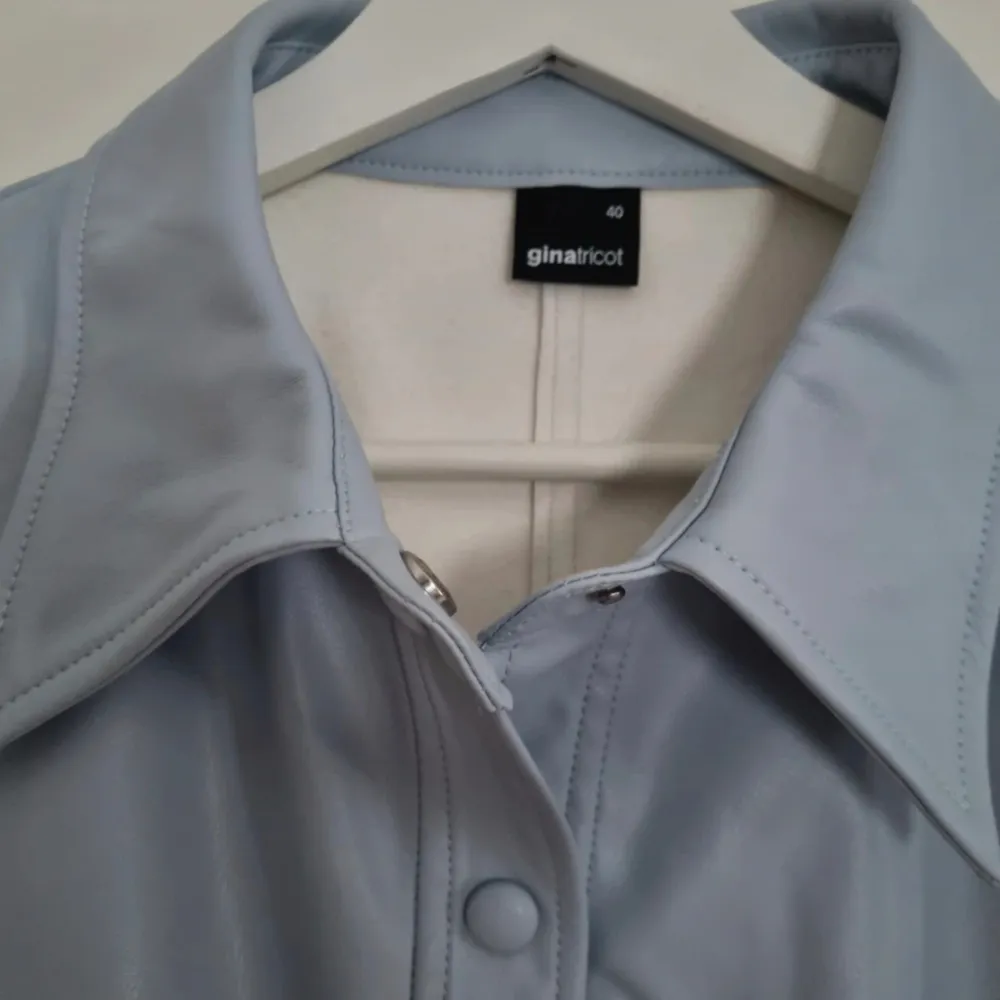 Ljusblå blus/skjorta faux leather, kort ärm, insytt rynk, knappar och krage. Storlek 40, bedömmer som normal (har lite stretch). Under arm-arm/byst ca 45 cm, midja ca 40 cm (x2 för omkrets). Längd ca 56 cm. Oanvänd, inga defekter, mjuk insida.. Skjortor.