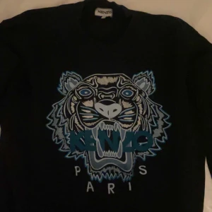 Svart tröja med tigertryck från Kenzo - Säljer en svart tröja från Kenzo med ett stort tigertryck i blått och vitt på framsidan. Tröjan har långa ärmar och en rund halsringning. Perfekt för en cool och avslappnad stil.