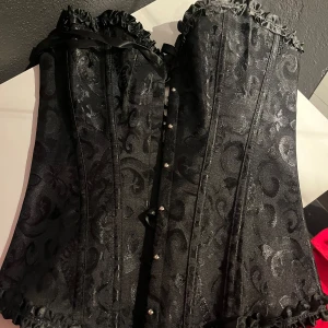 Svart korsett med broderat mönster - Elegant svart korsett med vackert broderat mönster och snörning bak. Den har dekorativa volanger upptill och nertill samt knäppning framtill. Perfekt för en stilfull look.