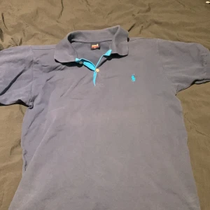 Blå pikétröja från Ralph Lauren - Säljer en klassisk blå pikétröja från Ralph Lauren med en ljusblå krage och knappslå. Tröjan har korta ärmar och den ikoniska loggan broderad på bröstet. Perfekt för en stilren look.