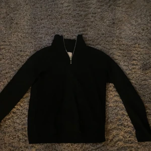 Svart stickad tröja med dragkedja - Säljer en stilren svart stickad tröja med dragkedja vid halsen. Perfekt för kyligare dagar och enkel att matcha med olika outfits. Tröjan har långa ärmar och en bekväm passform.