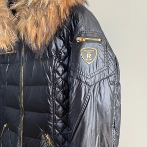 Svart dunjacka med äkta pälsdetalj från Rockandblue - Snygg svart dunjacka från Rockandblue i storlek 38, med en lyxig pälsdetalj runt kragen. Jackan har dragkedjor i guld och flera praktiska fickor. Perfekt för kyliga dagar med sin quiltade design och långa ärmar. Jackan är använd endast en gång 