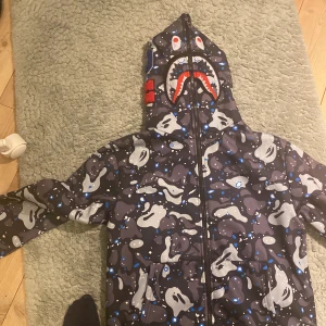 Camouflage hoodie med hajmotiv - Säljer en cool hoodie med ett unikt hajmotiv på huvan. Jackan har ett grått och svart camouflagemönster med blå detaljer. Den har dragkedja framtill och långa ärmar. Perfekt för dig som vill sticka ut!
