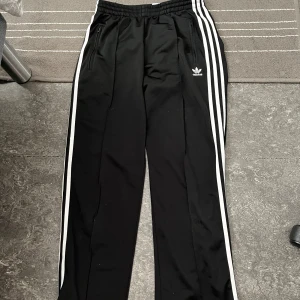 Svarta träningsbyxor från Adidas - Snygga svarta träningsbyxor från Adidas med de klassiska tre vita ränderna längs sidorna. Byxorna har en elastisk midja och dragkedjor vid fickorna för säker förvaring. Passar både storlek S & M