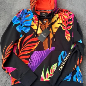 Färgglad hoodie från Carlo Colucci - Säljer en färgglad hoodie från Carlo Colucci med ett tropiskt mönster i blått, rosa, orange och lila. Hoodien har en stor ficka fram och märkesnamnet tryckt längs ärmen. Perfekt för att sticka ut i mängden!