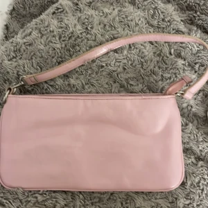 Rosa axelväska - Snygg rosa axelväska i glansigt material Den har en enkel design med en justerbar axelrem och dragkedja upptill. Perfekt för att bära med sig det nödvändigaste.