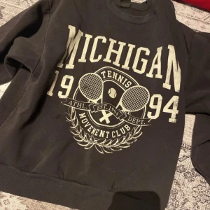 Svart sweatshirt från Gina Tricot - Säljer en svart sweatshirt från Gina Tricot med tryck av texten 'Michigan 1994' och tennismotiv. Tröjan har långa ärmar och ribbade muddar vid ärmslut och nederkant. Perfekt för en sportig look! Fläckarna går bort i tvätten