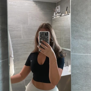 Svart kortärmad topp från ICANIWILL - Säljer en svart kortärmad topp från ICANIWILL i storlek XS. Toppen är tillverkad av återvunnen nylon och spandex, vilket ger en stretchig och bekväm passform. Den har en djup v-ringning och en liten logga på bröstet. Perfekt för en stilren och sportig look.