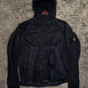 Svart vindjacka från Stone Island - Säljer en svart vindjacka från Stone Island med huva och dragkedja. Jackan har en ikonisk logga på ärmen och är perfekt för blåsiga dagar. Den är tillverkad i ett lätt syntetmaterial och har långa ärmar. Pris kan diskuteras!
