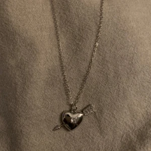 Silverfärgat halsband med hjärthänge - Charmigt silverfärgat halsband med ett hjärthänge genomborrat av en pil. Perfekt för att ge en romantisk touch till din outfit. Kedjan är tunn och elegant, vilket gör det lätt att bära till olika tillfällen.