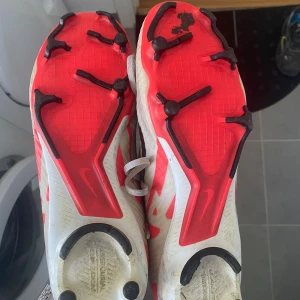 Nike fotbollsskor med snörning - Säljer ett par Nike fotbollsskor i vitt och rött med svarta detaljer. Skorna har snörning och en robust sula med dobbar för bra grepp på planen. Perfekta för fotbollsträning och matcher.