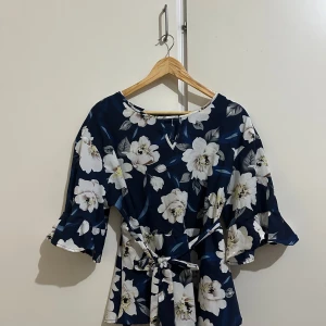 Blommig blus med volangärmar - Säljer en vacker blommig blus i mörkblått med stora vita blommor. Blusen har trekvartsärmar med volangdetaljer och ett knytband i midjan för en smickrande passform. Perfekt för vår och sommar!