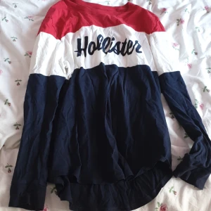 Randig långärmad tröja från Hollister - Säljer en cool långärmad tröja från Hollister med ränder i rött, vitt och blått. Tröjan har en avslappnad passform och är perfekt för en casual look. Hollister-loggan är tryckt på bröstet.