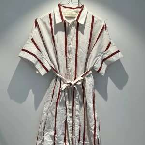 Vit och röd randig skjorta från By Malene Birger - Snygg vit skjorta med röda ränder från By Malene Birger. Den har korta ärmar och en klassisk krage. Skjortan kommer med ett knytband i midjan för en fin passform. 