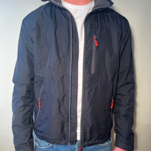 Jacka från Helly Hansen - Säljer en blå vindjacka från Helly Hansen med röda dragkedjor och HH-logga på ärmen. Jackan har en hög krage och är perfekt för blåsiga dagar. Den är tillverkad i ett slitstarkt material och har flera praktiska fickor med dragkedja.