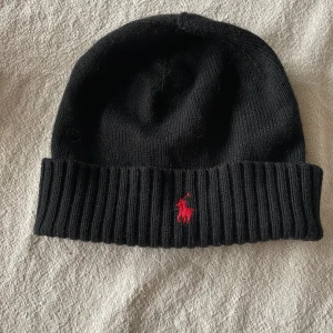 Svart mössa från Polo Ralph Lauren - Snygg svart mössa från Polo Ralph Lauren med ribbad kant och röd broderad logga framtill. Perfekt för att hålla värmen under kyliga dagar.