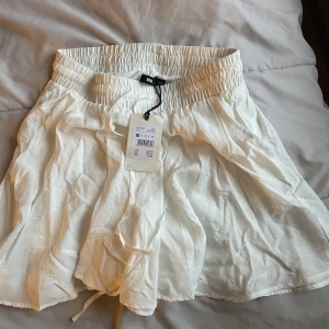 Vit kjol - Säljer en vit kjol med elastisk midja och volangdetaljer. Kjolen är kort och har en luftig passform, perfekt för varma dagar. Den är gjord i ett lätt och bekvämt material.