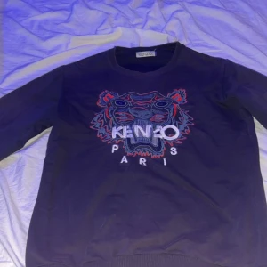 Mörkblå sweatshirt från Kenzo - Säljer en mörkblå sweatshirt från Kenzo med ett broderat tigerhuvud och texten 'Kenzo Paris' på framsidan. Tröjan har långa ärmar och en rund halsringning. Perfekt för en stilren och avslappnad look.( den passar S men den är L i junior storlek) Vet ej om den är äkta eller fejk där av priset) kan mötas i jkpg eller bara frakta. Mvh Affe 